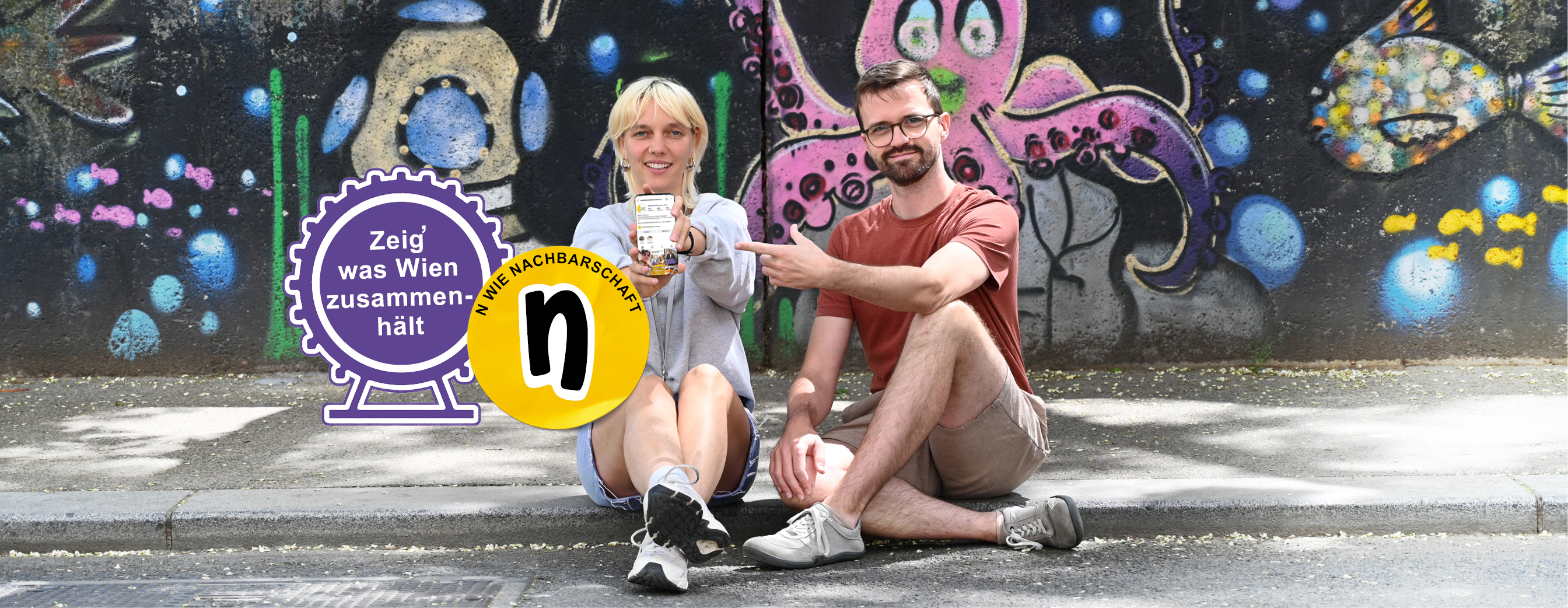 Eine Frau und ein Mann halten ein Smartphone ins Bild, auf dem das Projekt "WIEnIEMAN ALLEIN" zu sehen ist
