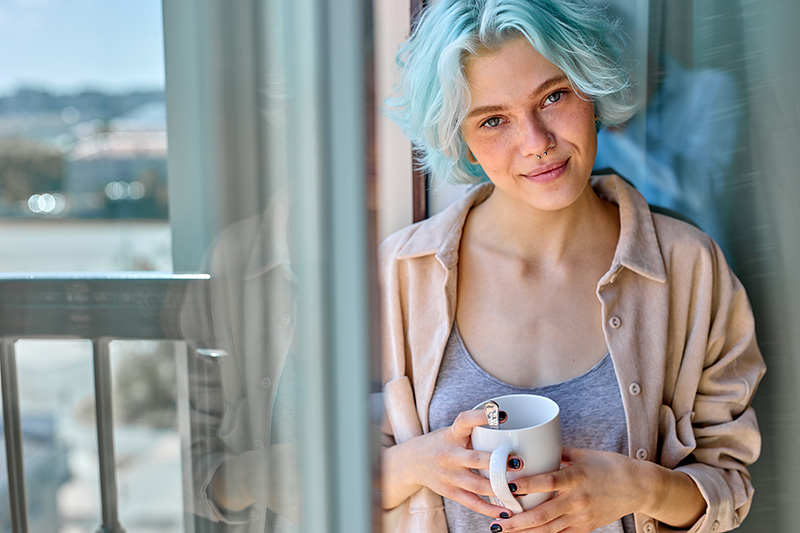 Eine junge Frau steht beim Fenster mit einer Tasse in der Hand