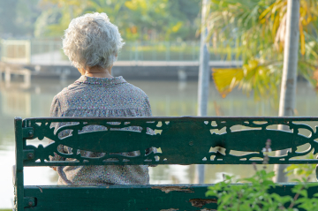 Eine ältere Frau sitzt alleine auf einer Parkbank an einem Teich.