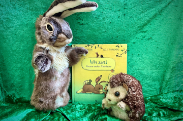Zwei Kuscheltiere - ein Hase und ein Igel, dahinter das Bilderbuch "Wir zwei" stehend